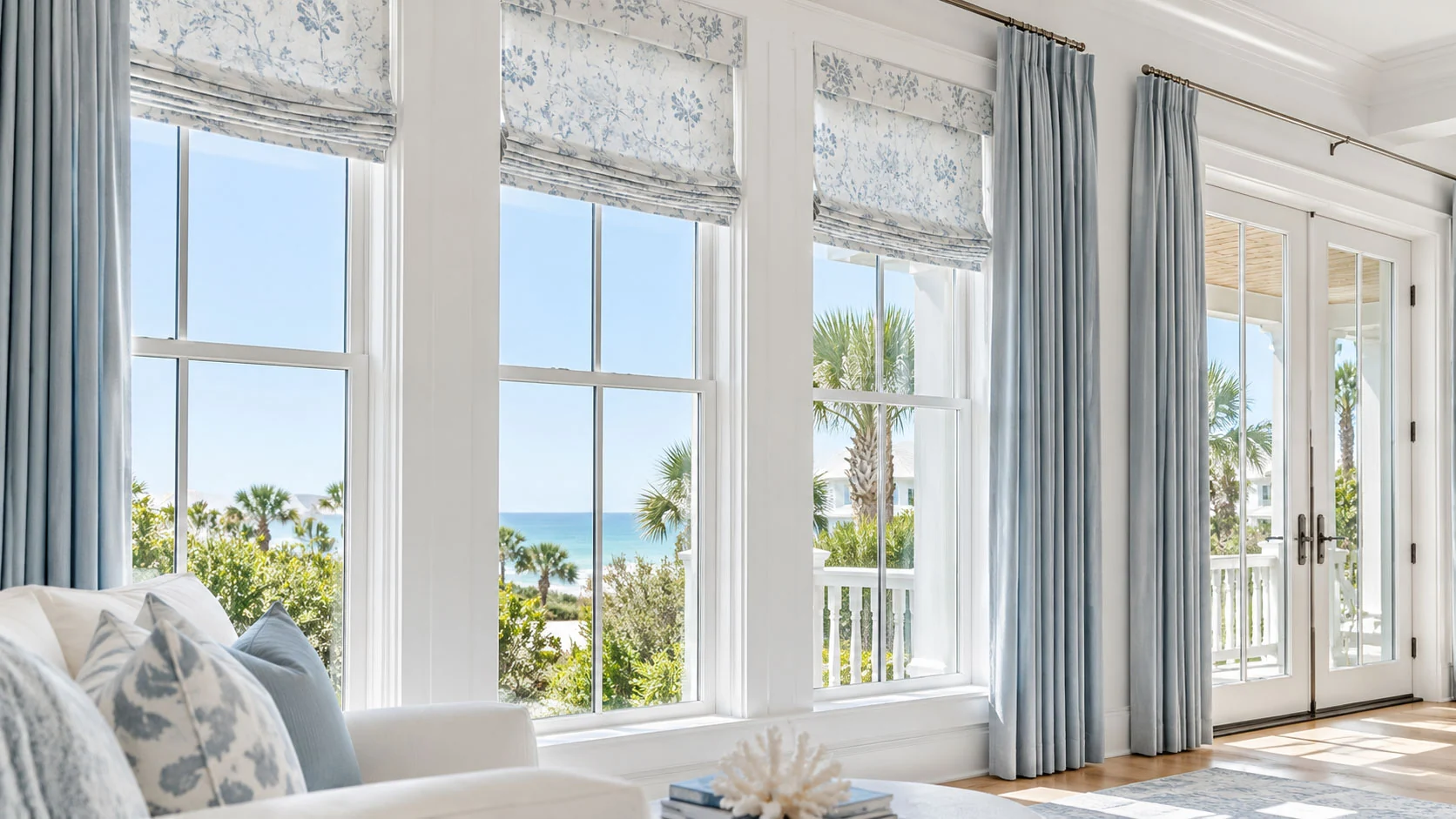 Floral Roman shades and pale blue drapery beside sunlit coastal windows and French doors