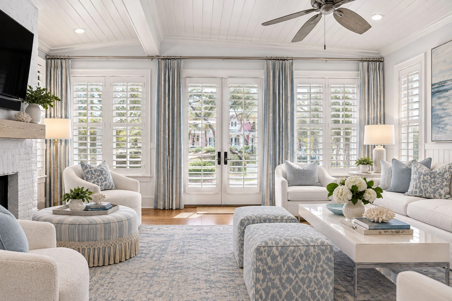 Pale-blue drapery and white plantation shutters frame French doors in a layered coastal sitting room