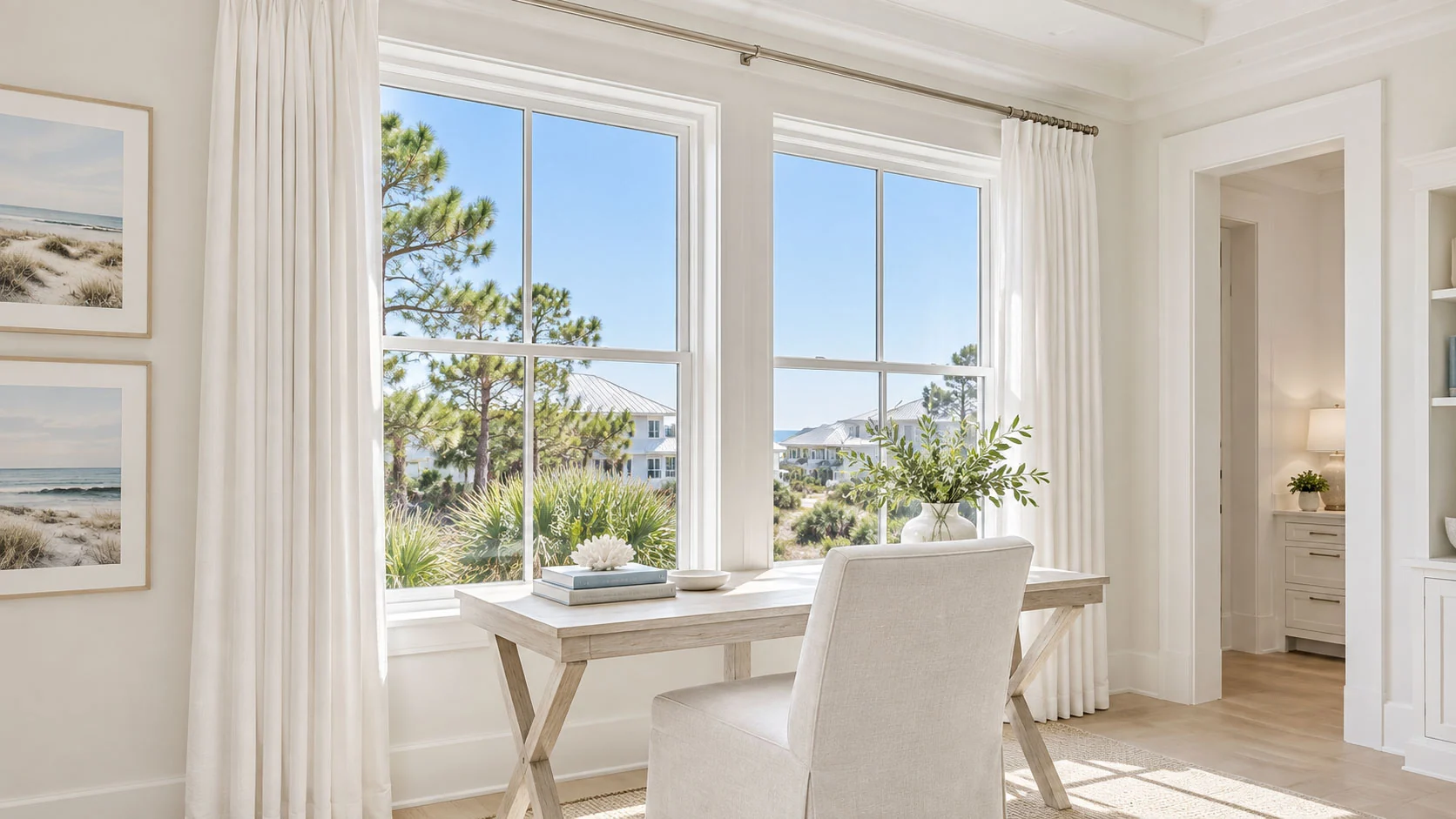 Full-length white pleated drapery frames two large windows above a pale wood desk in a sunlit coastal study