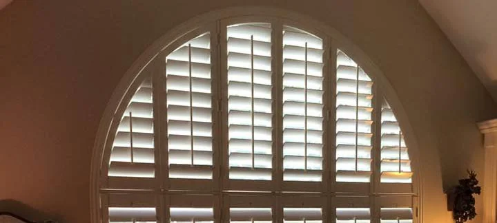 White plantation shutters fitted to a large arched interior window in a warmly lit room.